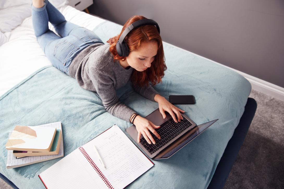 Regardant vers le bas sur l'étudiant féminin d'université utilisant des écouteurs se trouvant sur le lit fonctionnant sur l'ordinateur portatif